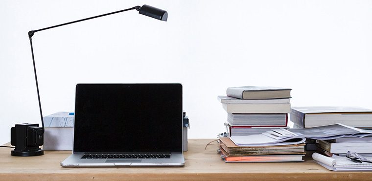 Lamp and open laptop with a dark screen on a desk with a stack of books on the right.